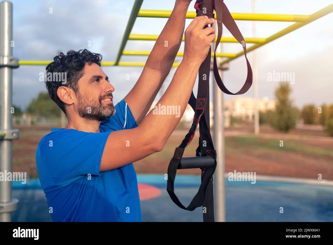 Caucasian man preparing a great TRX workout at outside in sunny park. Young handsome guy in sportswear doing exercising outdoors. Healthy lifestyle co Stock Photo