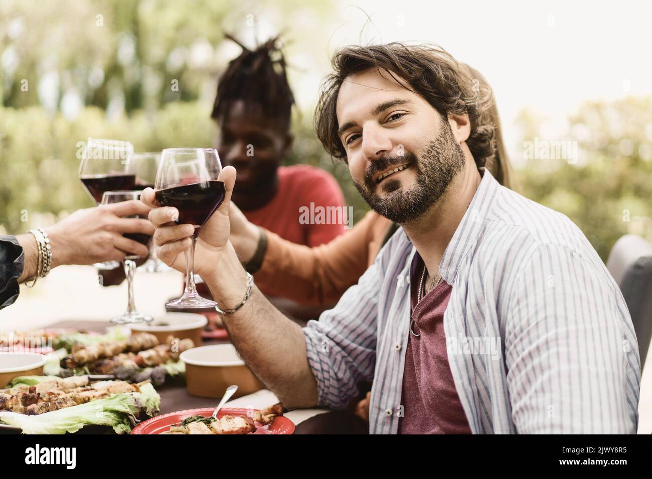 Spending time with friends. Handsome bearded forty man toasting with red wine and smiling while sitting with his friends in the farmhouse restaurant c Stock Photo