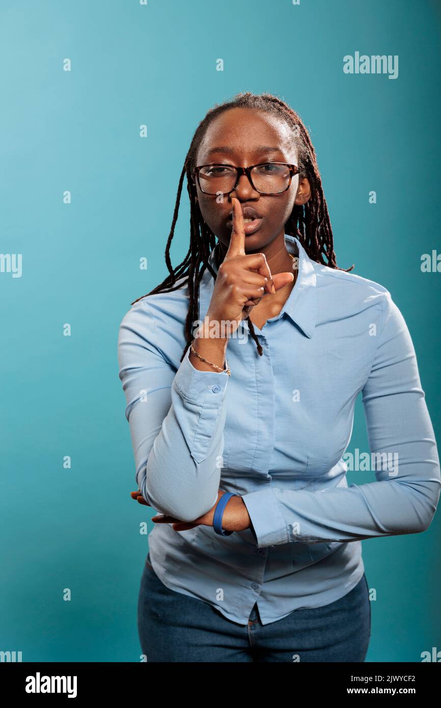 Attractive serious woman making silence gesture by putting forefinger over lips on blue background. Young adult person shushing people indicating secrecy and confidentiality. Stock Photo