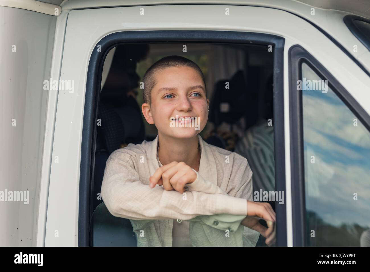 Young caucasian beautiful girl with shaved head looking out of the open window of a camping van in awe, marveling over the beautiful scenery. High quality photo Stock Photo