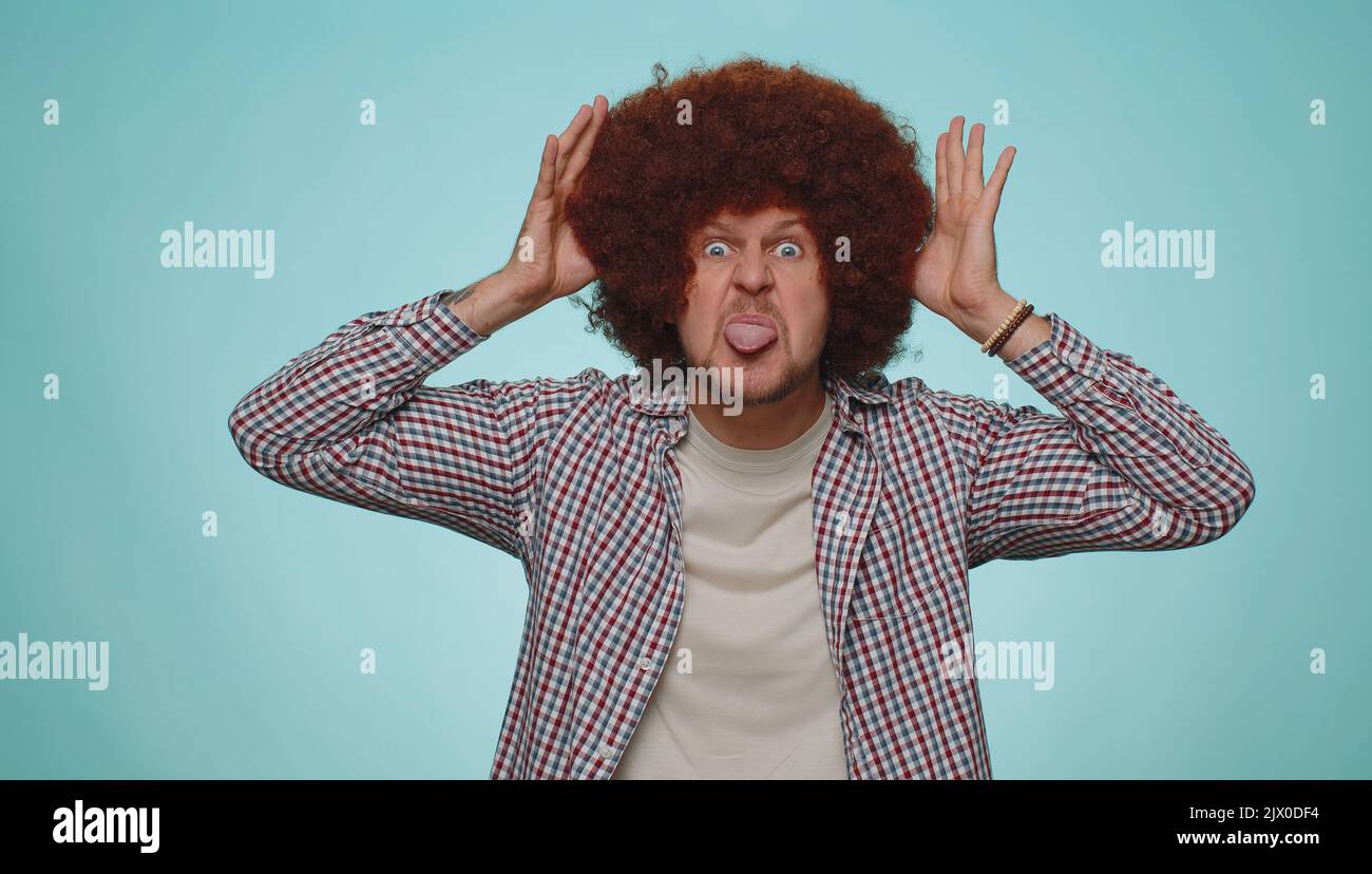 Cheerful funny man with lush hairstyle showing tongue making faces at camera, fooling around, joking, aping with silly face, teasing. Young adult guy boy isolated alone on blue studio background Stock Photo