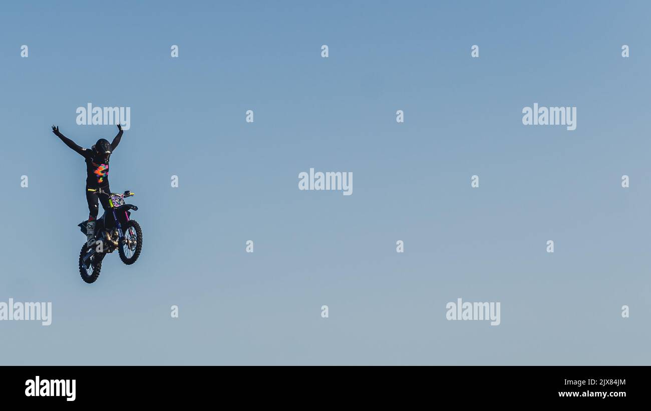 Wide panoramic shot of an extreme motorbike jump high in the air. Bike driver in protective gear standing on the bike and stretching arms in the air. Blue sky as a uniform seamless background. . High quality photo Stock Photo