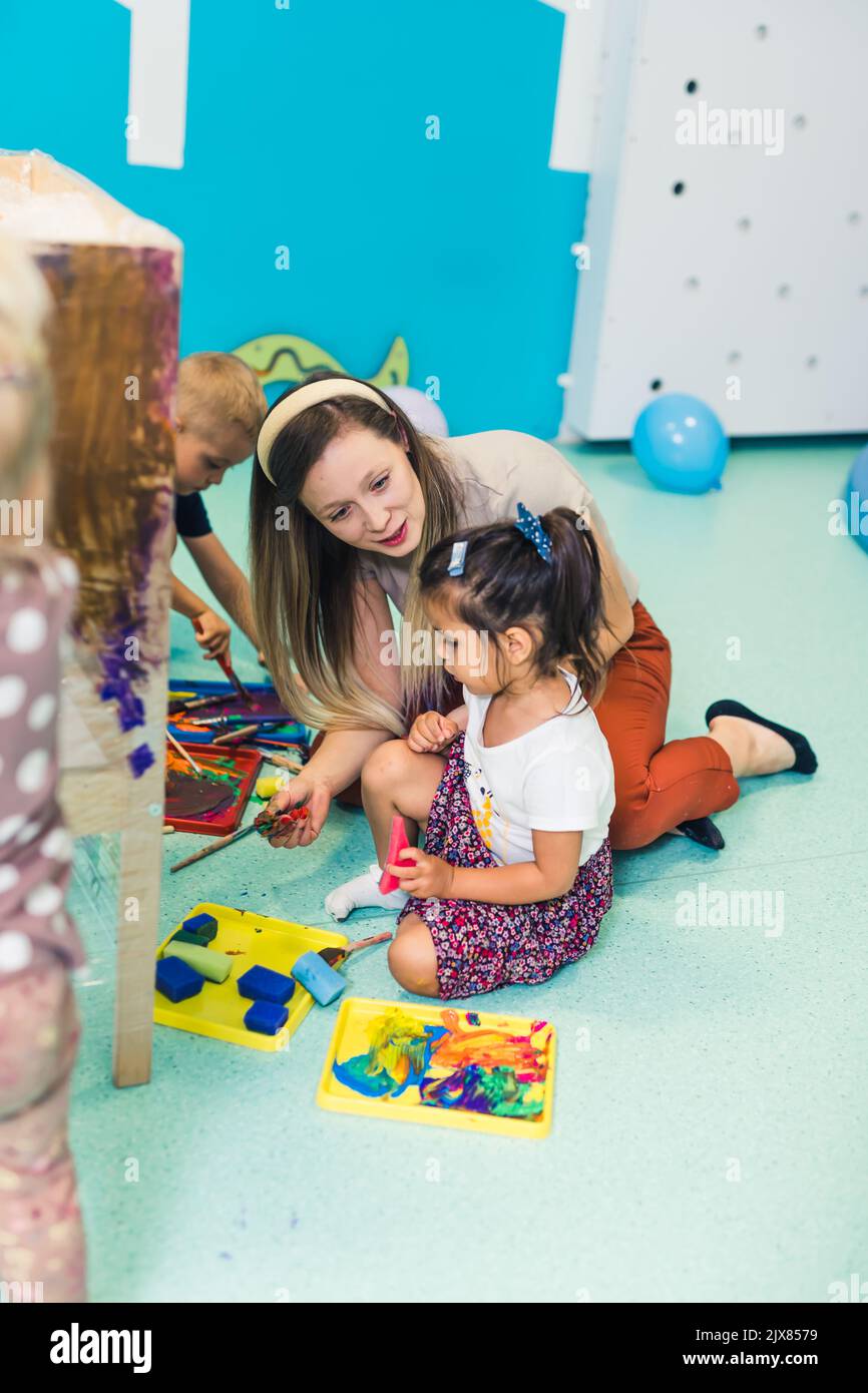 Cling film painting. Toddler painting with a sponge, brushes and paints on a cling film wrapped all the way round the wooden shelf unit. A teacher helping them. Creative activity for kids sensory skills development at the nursery school. High quality photo Stock Photo