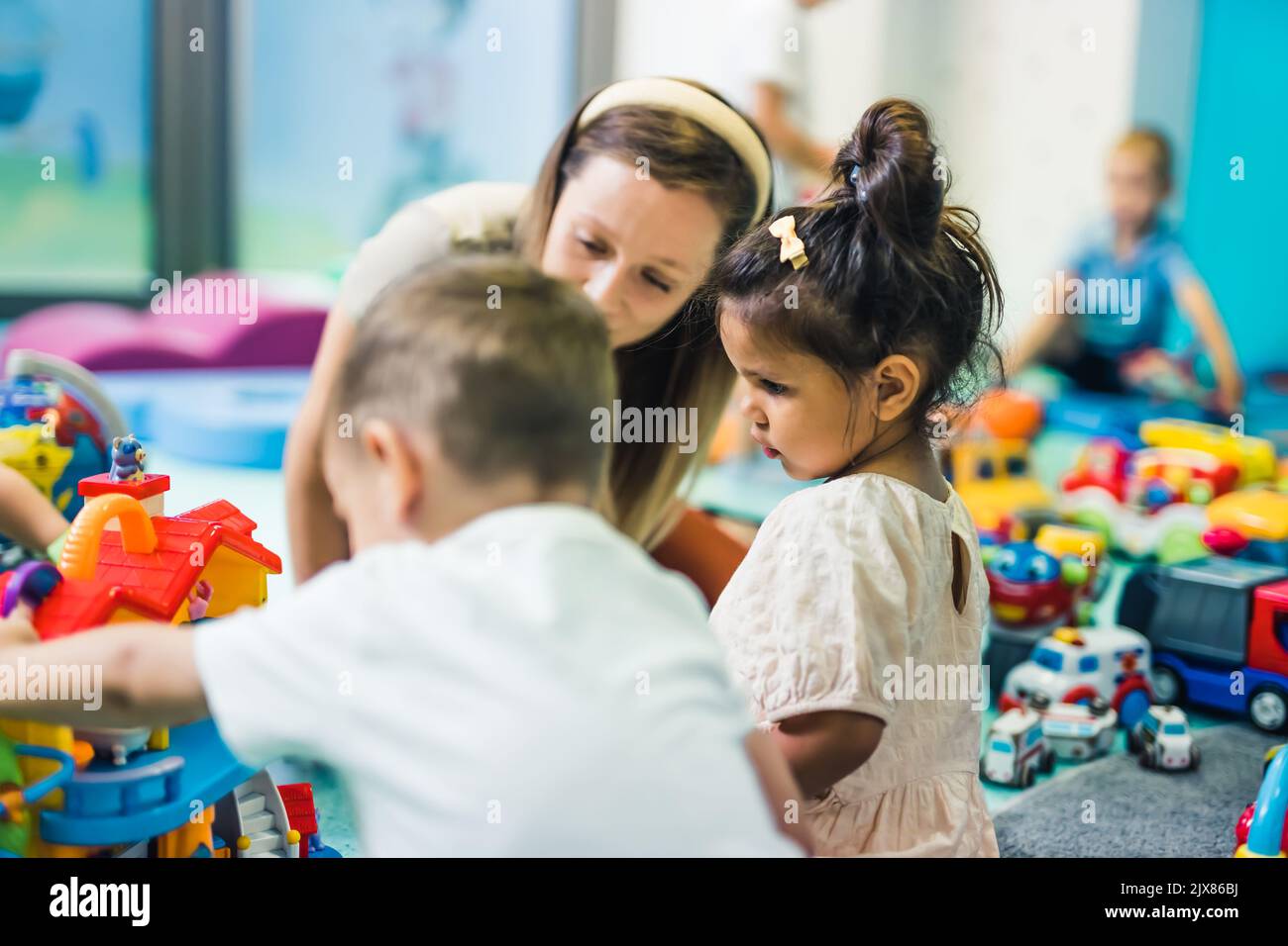 Toddlers and their nursery teacher playing with plastic building blocks and colorful car toys while sitting on the floor in a playroom. Concentration, fine motor and gross motor skills development, . High quality photo Stock Photo