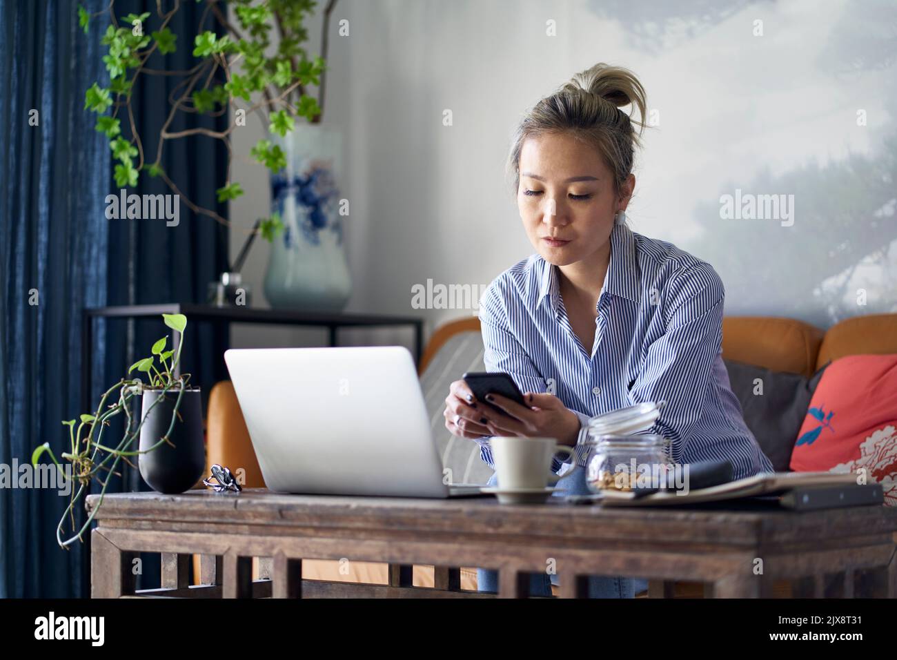 mature professional asian woman sitting in couch working from home using mobile phone and notebook computer Stock Photo