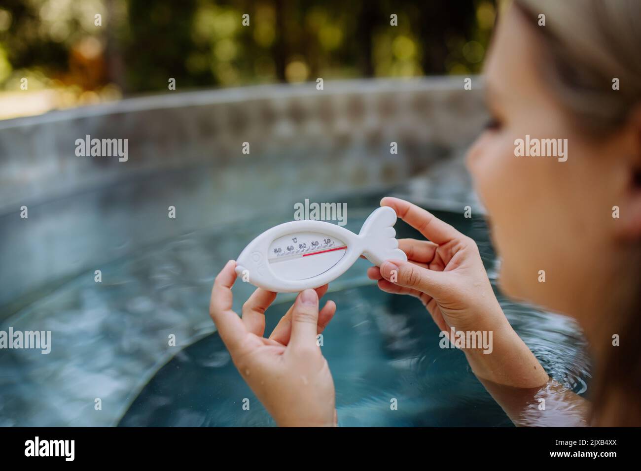 Young woman in bathtub, checking temperature of water with thermometer, ready for home spa procedure in hot tub outdoors. Wellness, body care, hygiene Stock Photo