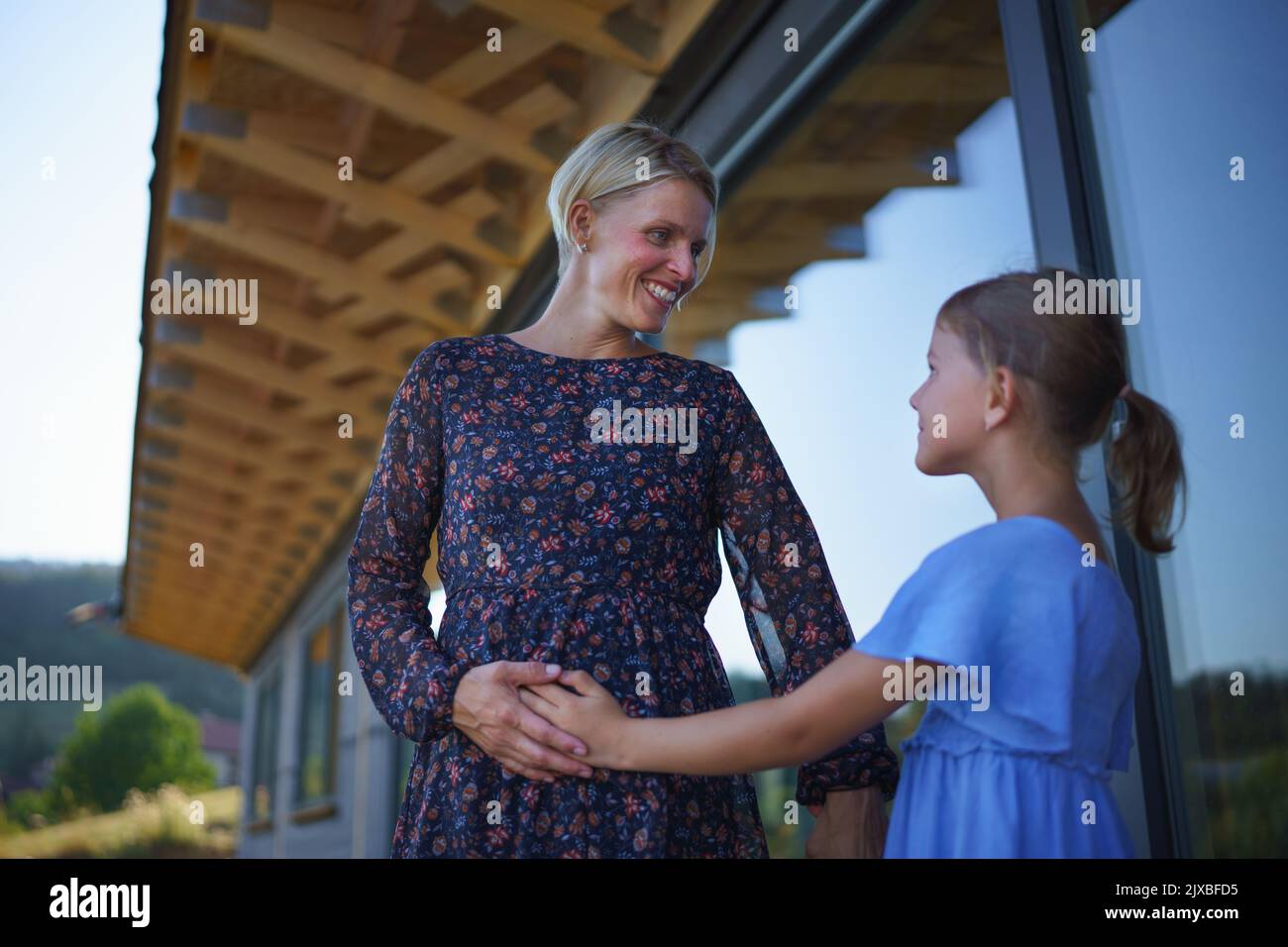 Little daughter stroking pregnant belly his mother, in front of their new wooden house. Stock Photo