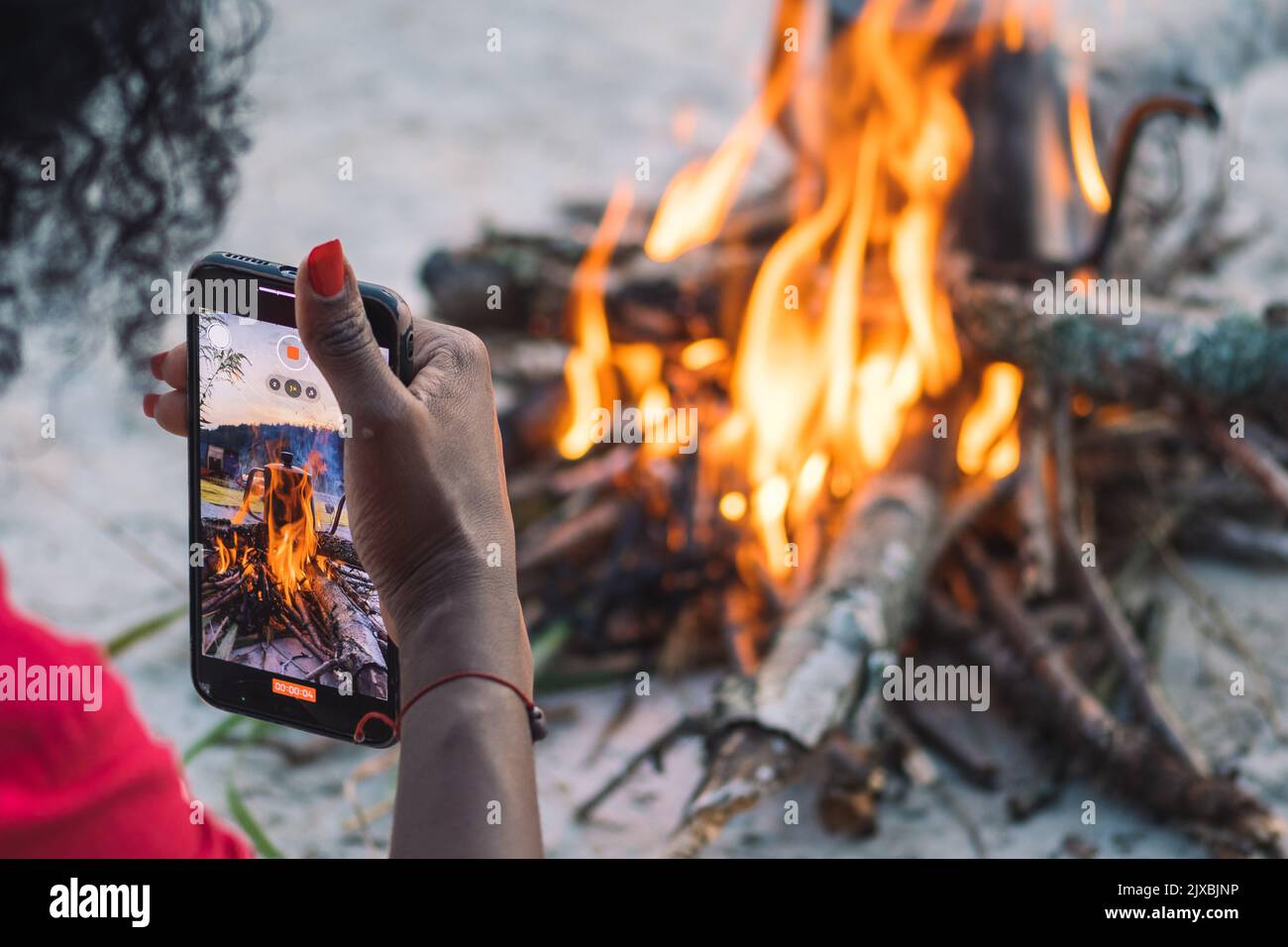 Girl taking photo or video with smartphone while preparing coffee or tea on a bonfire with branches and flames outdoor in the nature Stock Photo