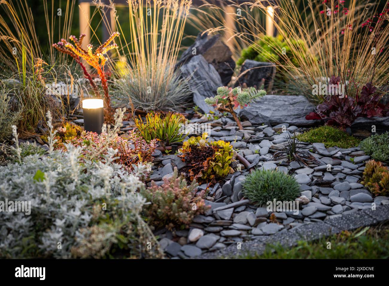 Beautifully Landscaped Flowerbed in Backyard Garden Decorated with Different Plants, Dark Pebbles and Outdoor Bollard Lighting Lamp. Landscaping Desig Stock Photo