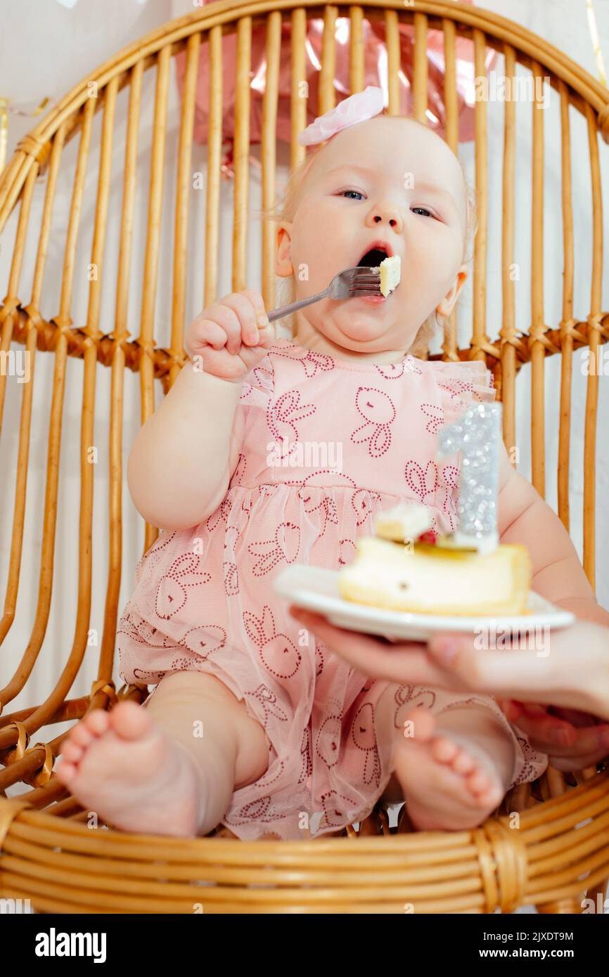 Vertical barefoot one year toddle kid in pink dress sitting on webbed chair and eating with joy piece of birthday cake Stock Photo