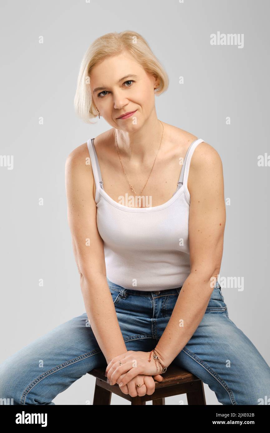 Barefoot senior woman in jeans and tank top sitting on tall chair in studio Stock Photo