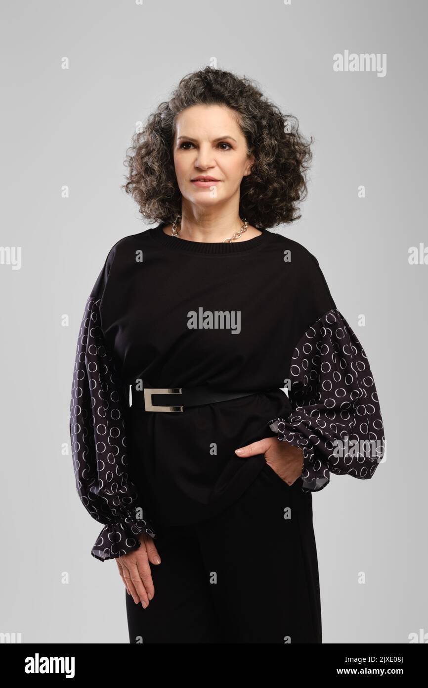 Senior woman with lush curly hair and black pantsuit posing in studio over grey background Stock Photo