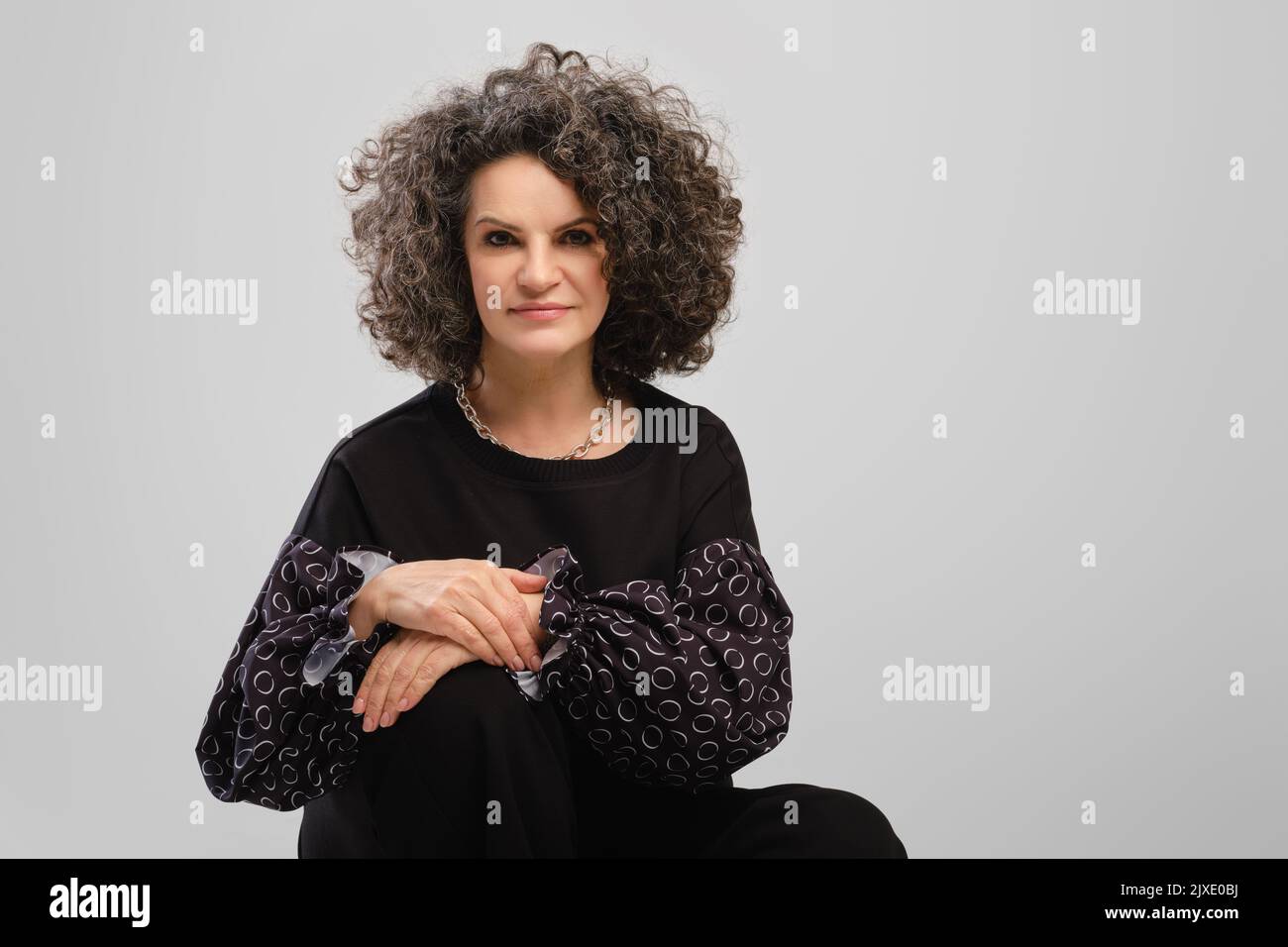 Portrait of senior woman with lush curly hair and black pantsuit posing in studio over grey background Stock Photo