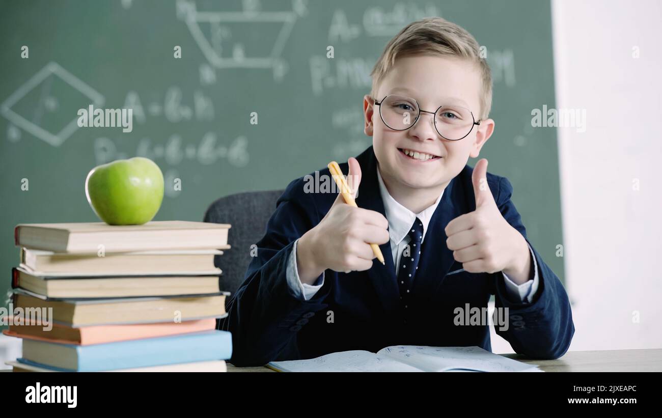 pleased schoolboy in glasses showing thumbs up near apple and books in ...