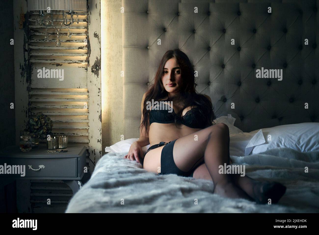Glamorous long-haired brunette young woman wearing black lingerie sits on a bed in a hotel room.  MODEL RELEASED Stock Photo