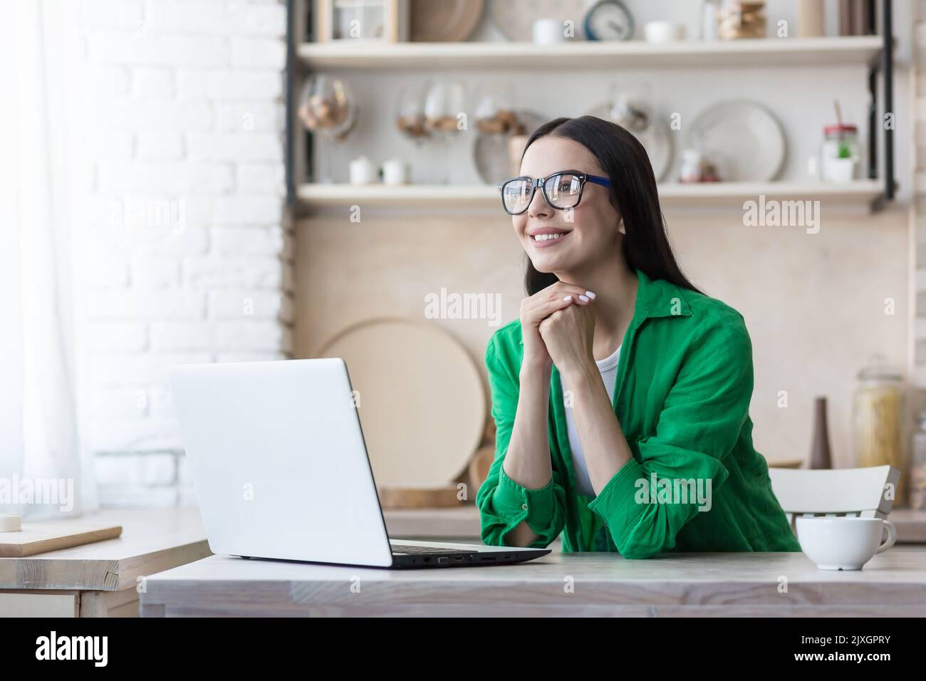 Blogging. Freelancer. A young woman in glasses sitting at home in the kitchen with a laptop, writing a post, working on social networks, chatting. She folded her hands under chin, thinking, smiling. Stock Photo