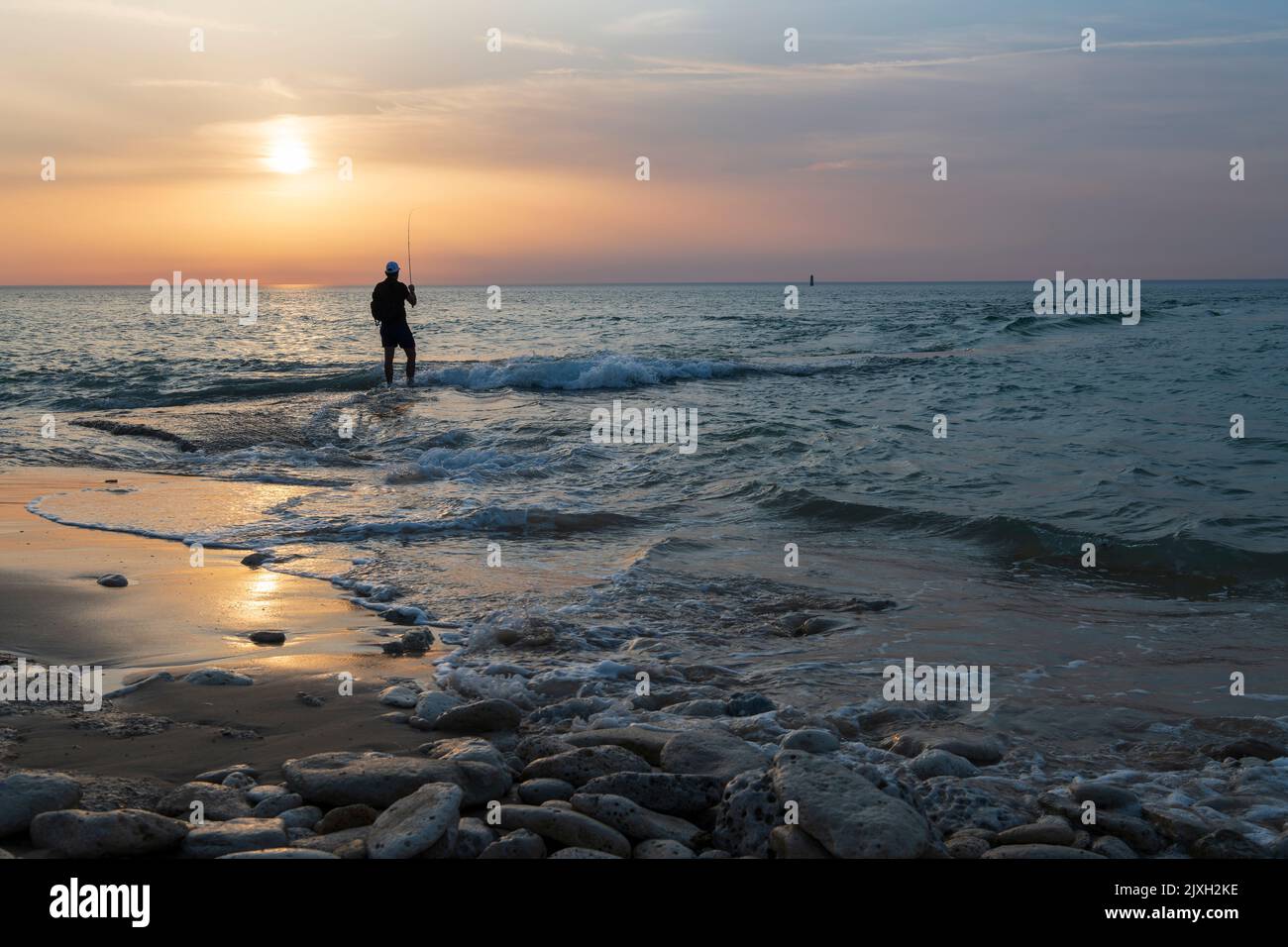 Young man fishing in the sunset by the shore with a breathtaking view. Stock Photo