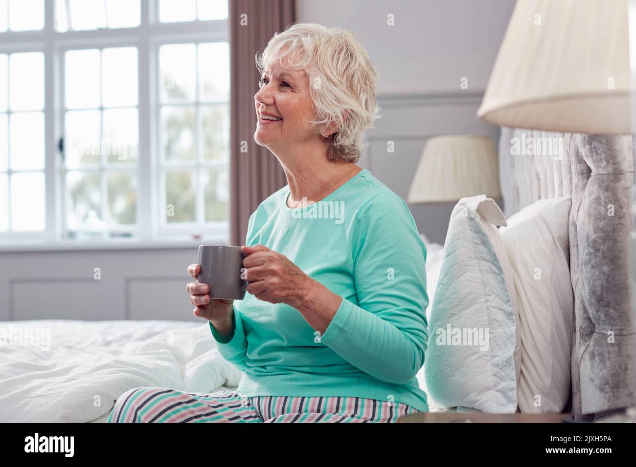 Senior Woman At Home Wearing Pyjamas Sitting On Bed Having Morning Cup Of Tea Stock Photo