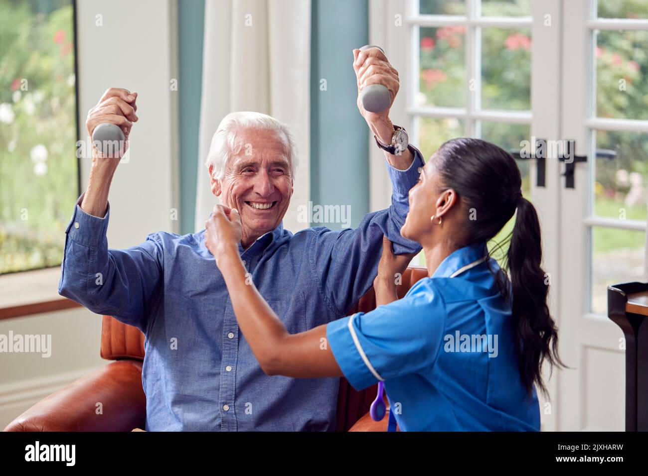 Female Nurse Or Physiotherapist In Uniform Helping Senior Man To Lift Hand Weights At Home Stock Photo