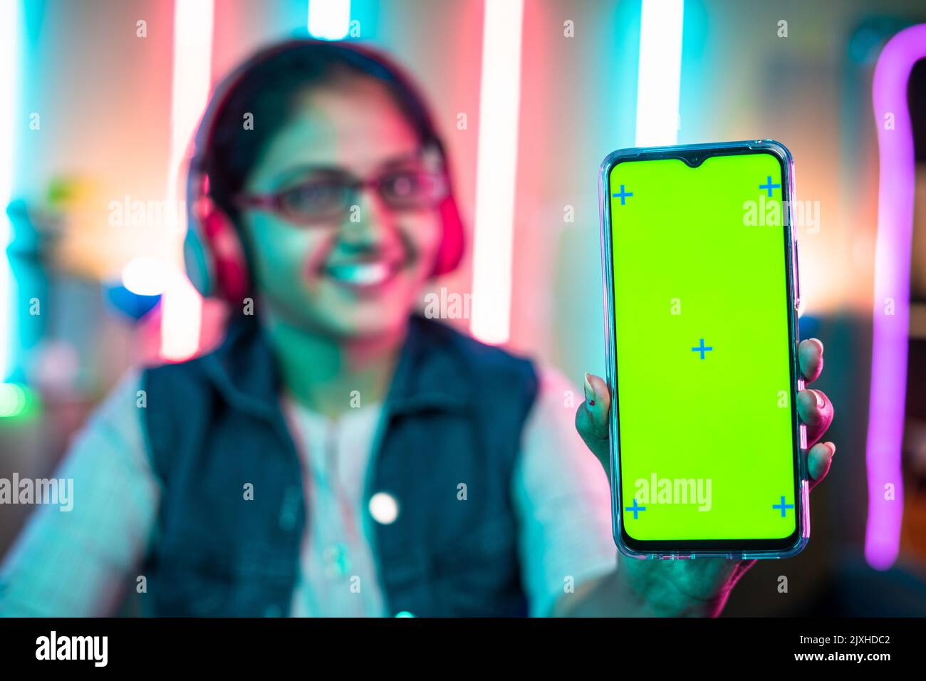 focus on mobile phone, Smiling girl showing green screen mobile phone by looking camera on neon gaming background. - concept of gaming advertisement Stock Photo