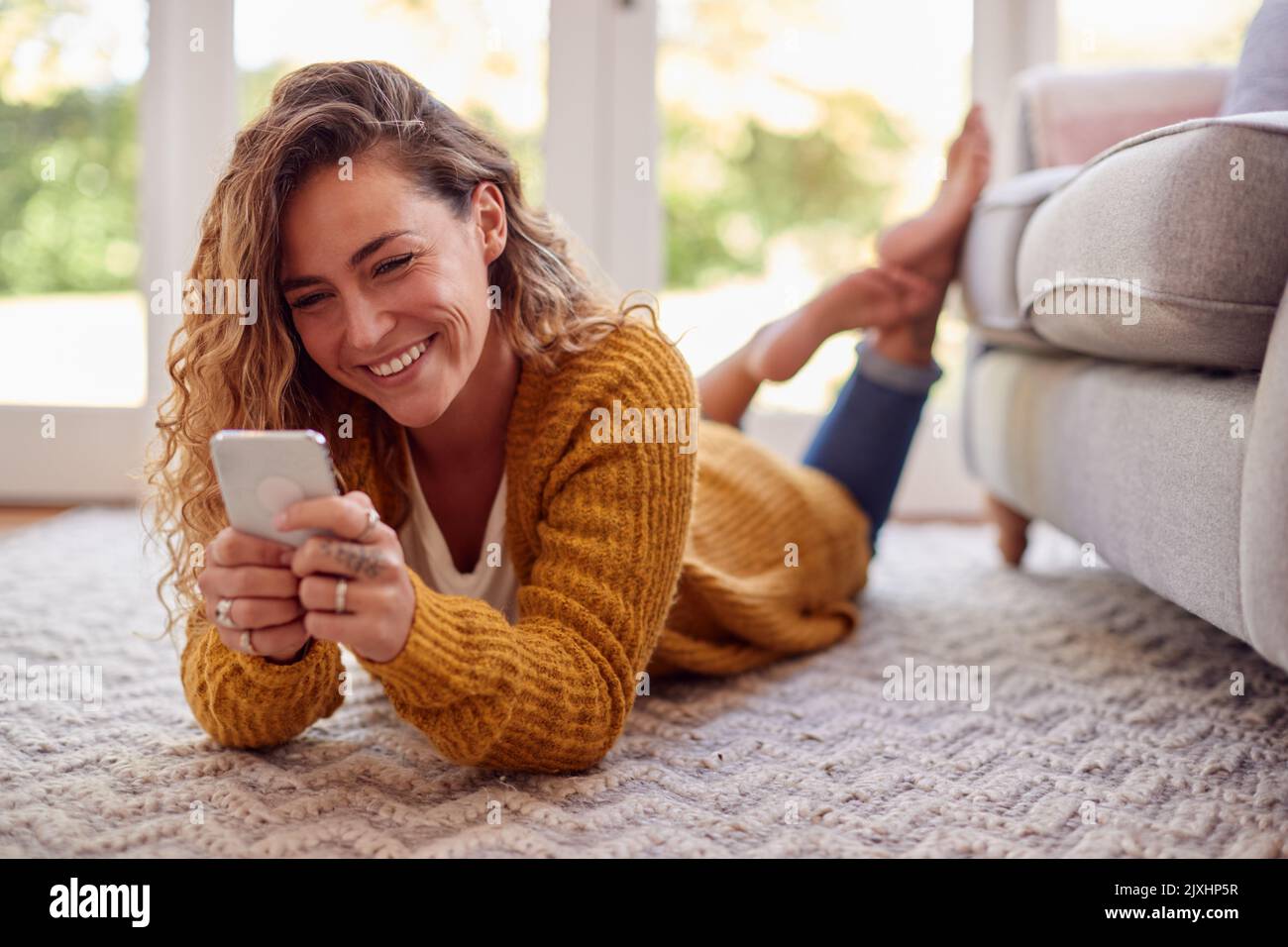 Woman In Warm Jumper Lying On Floor At Home Using Mobile Phone Stock Photo