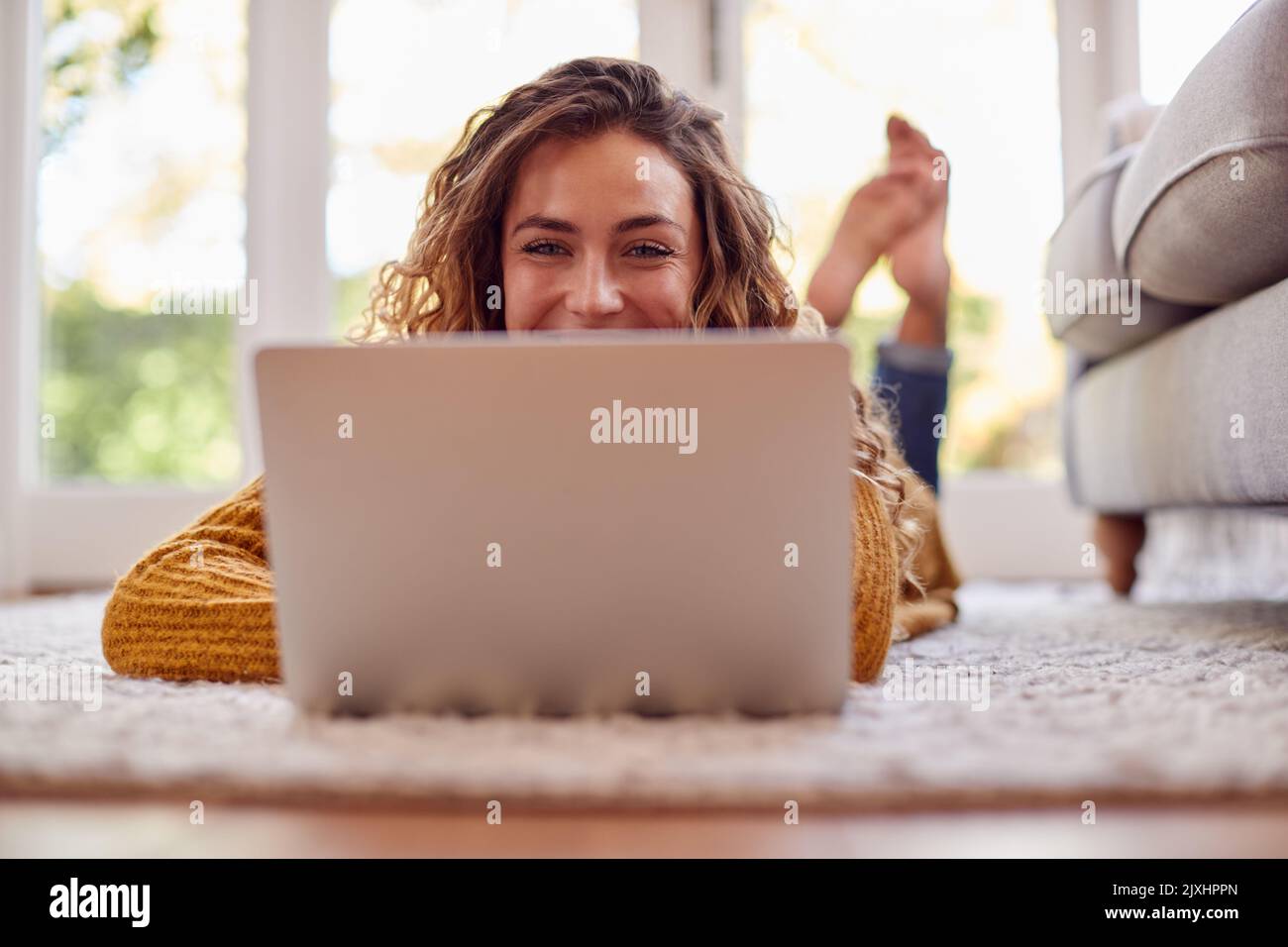 Woman In Warm Jumper Lying On Floor At Home Using Laptop Stock Photo