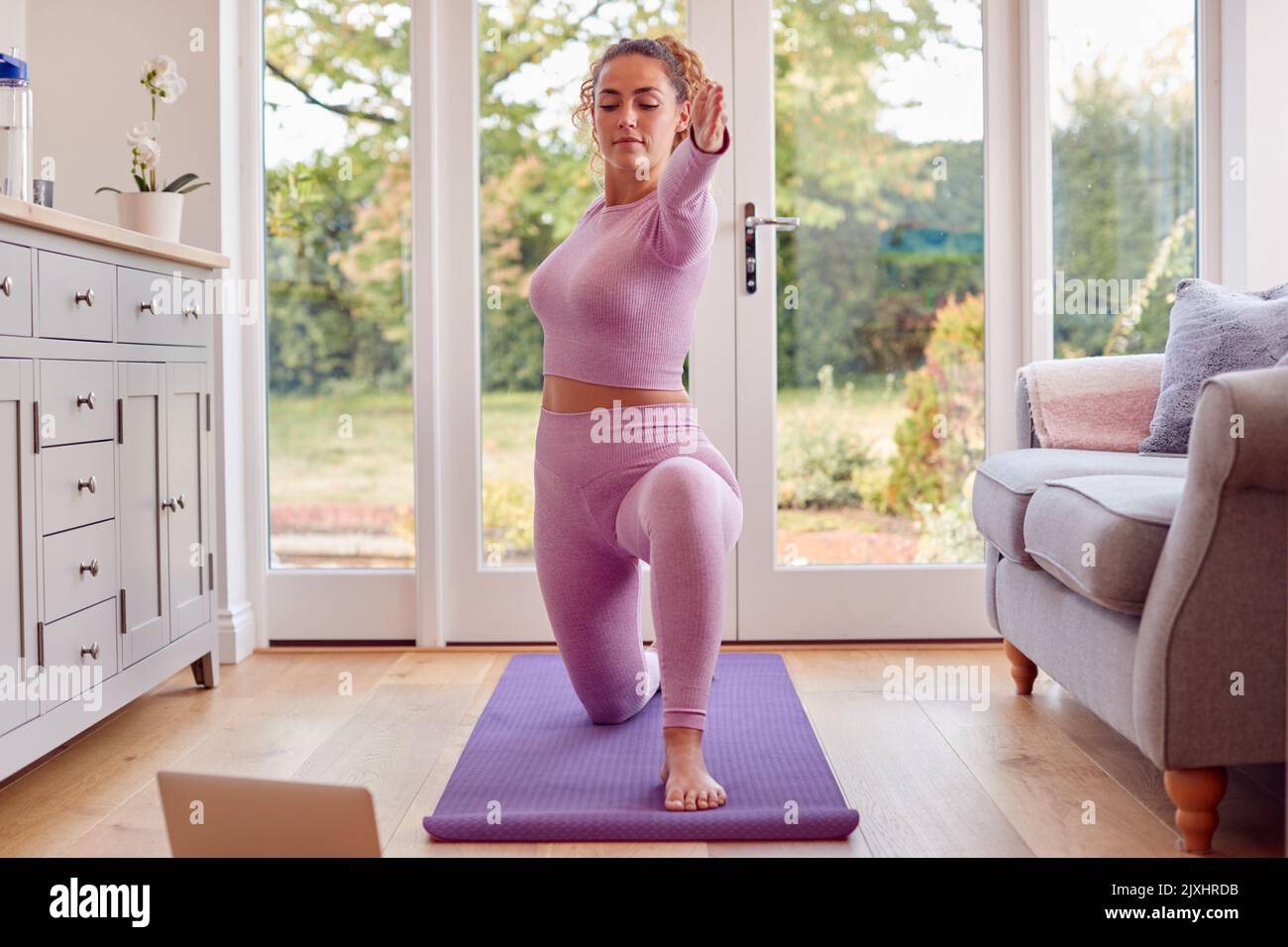 Young Woman Stretching On Mat At Home With Laptop Doing Online Yoga Class Stock Photo