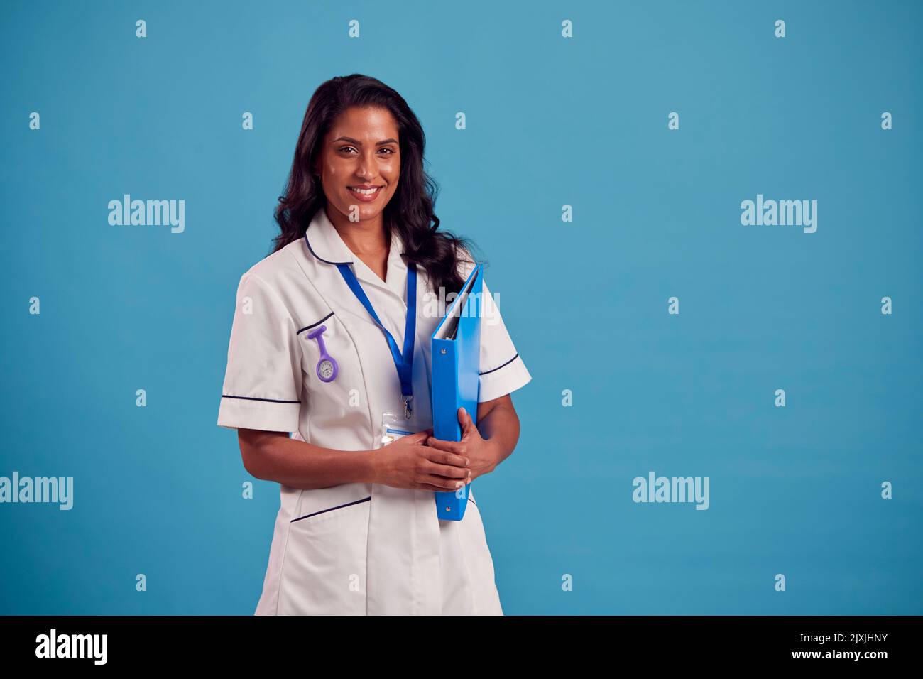 Portrait Of Smiling Female Mature Nurse Wearing Uniform Standing In Front Of Blue Studio Background Stock Photo