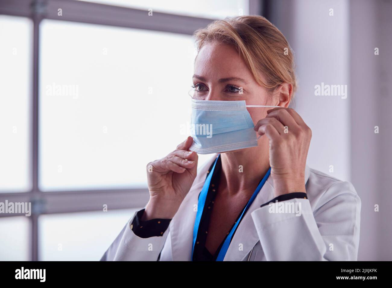 Mature Female Doctor Wearing White Coat Putting On Face Mask In
