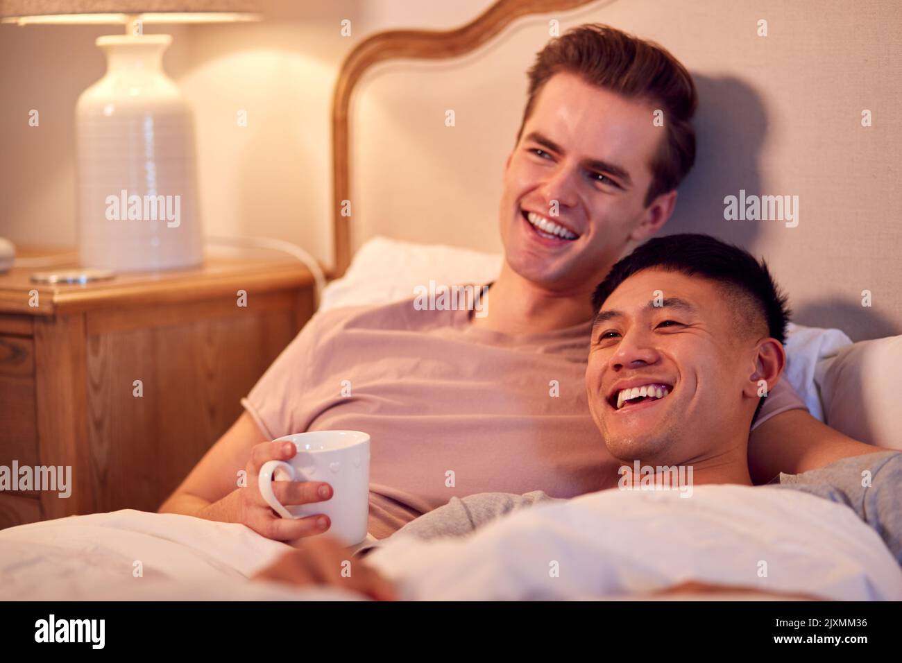 Loving Same Sex Male Couple Lying In Bed At Home Watching TV Together Stock Photo