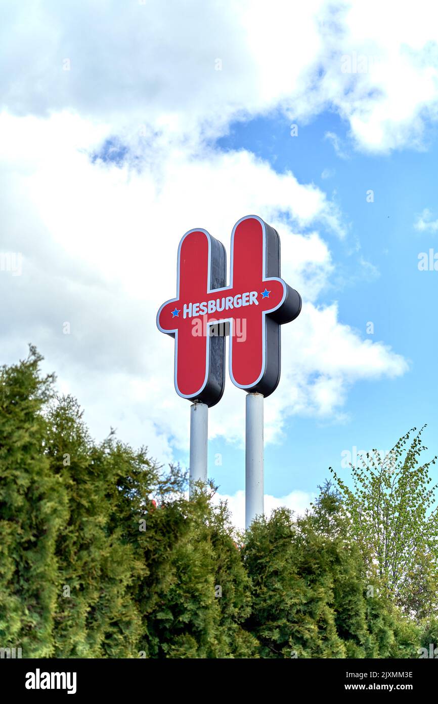 RIGA, LATVIA - MAY 13, 2022: Red Hesburger restaurant logo against blue cloudy sky during sunny day. Green coniferous trees below. Stock Photo