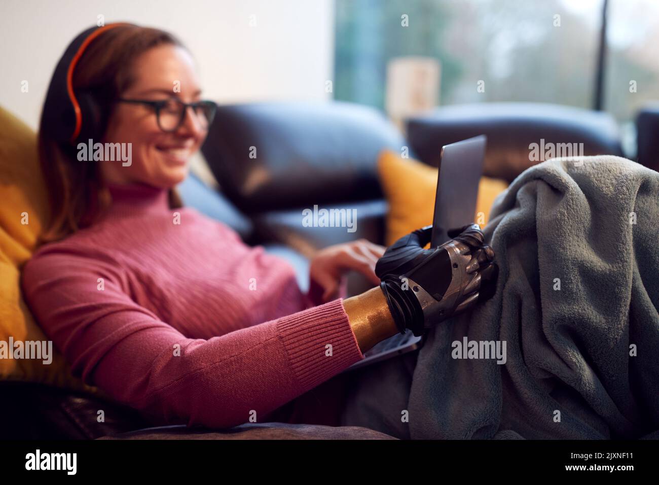 Woman With Prosthetic Arm Wearing Wireless Headphones And Working On Laptop On Sofa At Home Stock Photo