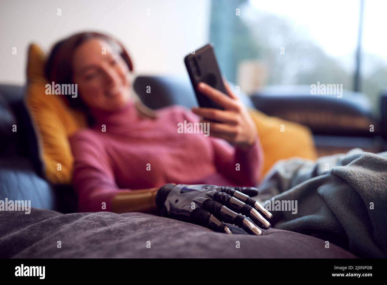 Woman With Prosthetic Arm Wearing Wireless Headphones Listening To Music On Mobile Phone On Sofa Stock Photo