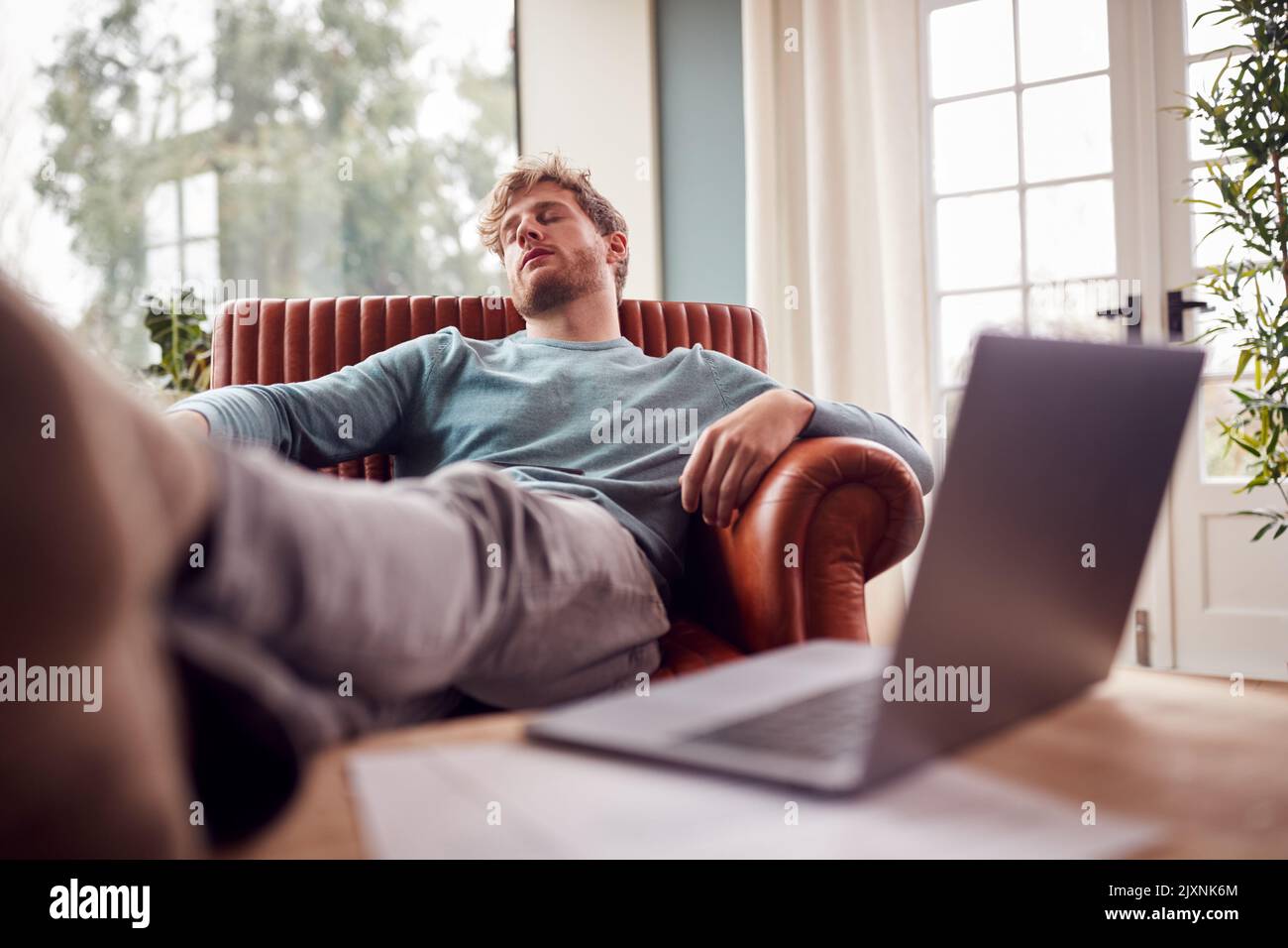 Tired Man Sleeping In Armchair With Feet Up At Home With Laptop In