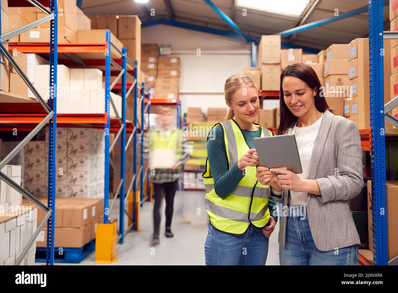 Manager Using Digital Tablet With Female Intern Inside Busy Warehouse Facility Stock Photo