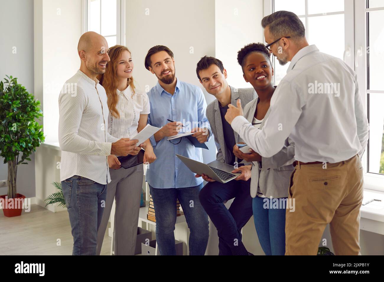 Friendly male business leader or trainer is talking to group of people standing in office near him Stock Photo