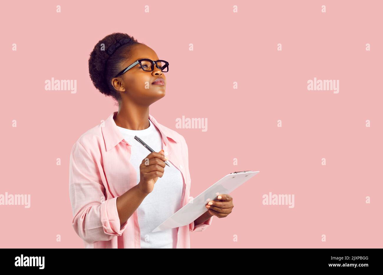 Young African American woman student wearing glasses holds clipboard stands in pink studio Stock Photo