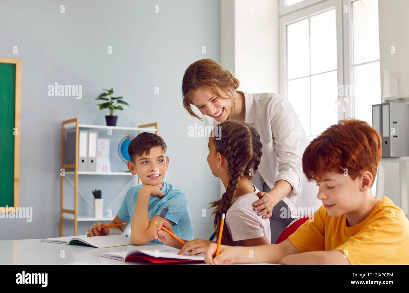 Friendly smiling female teacher helping her little students during lesson in classroom. Stock Photo