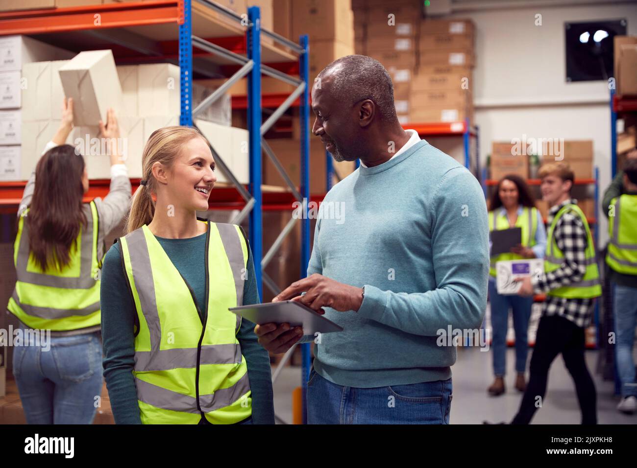 Female Intern With Team Leader Looking At Digital Tablet Inside Busy Warehouse Facility Stock Photo