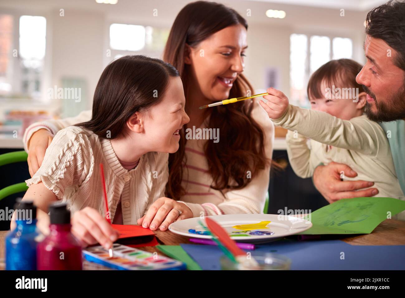 Family With Down Syndrome Daughter Sitting Around Table At Home Doing Craft Together Stock Photo