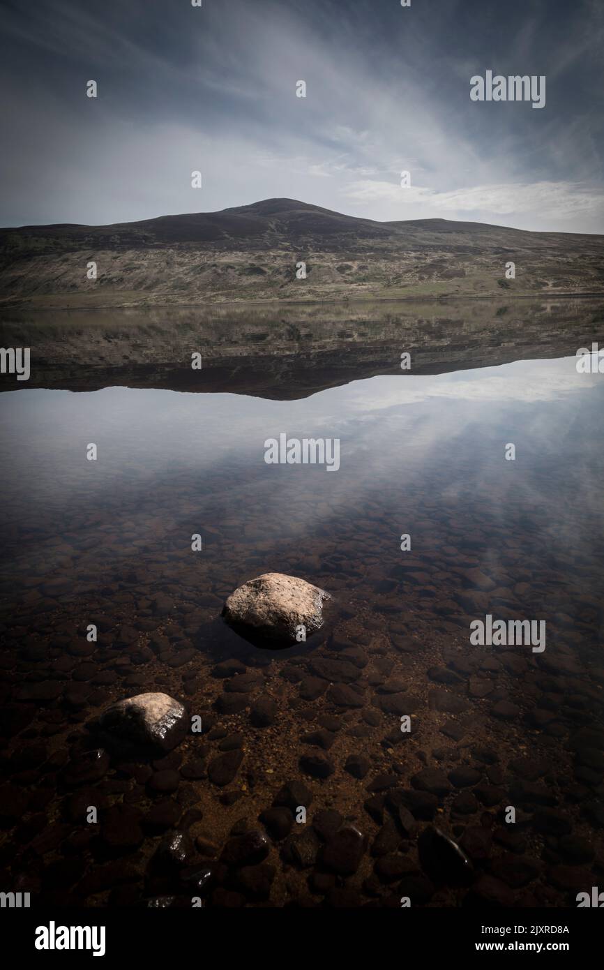 Rocks protrude from the still clear water of Loch Loyal, reflecting the mountain of Beinn Stumanadh. Stock Photo