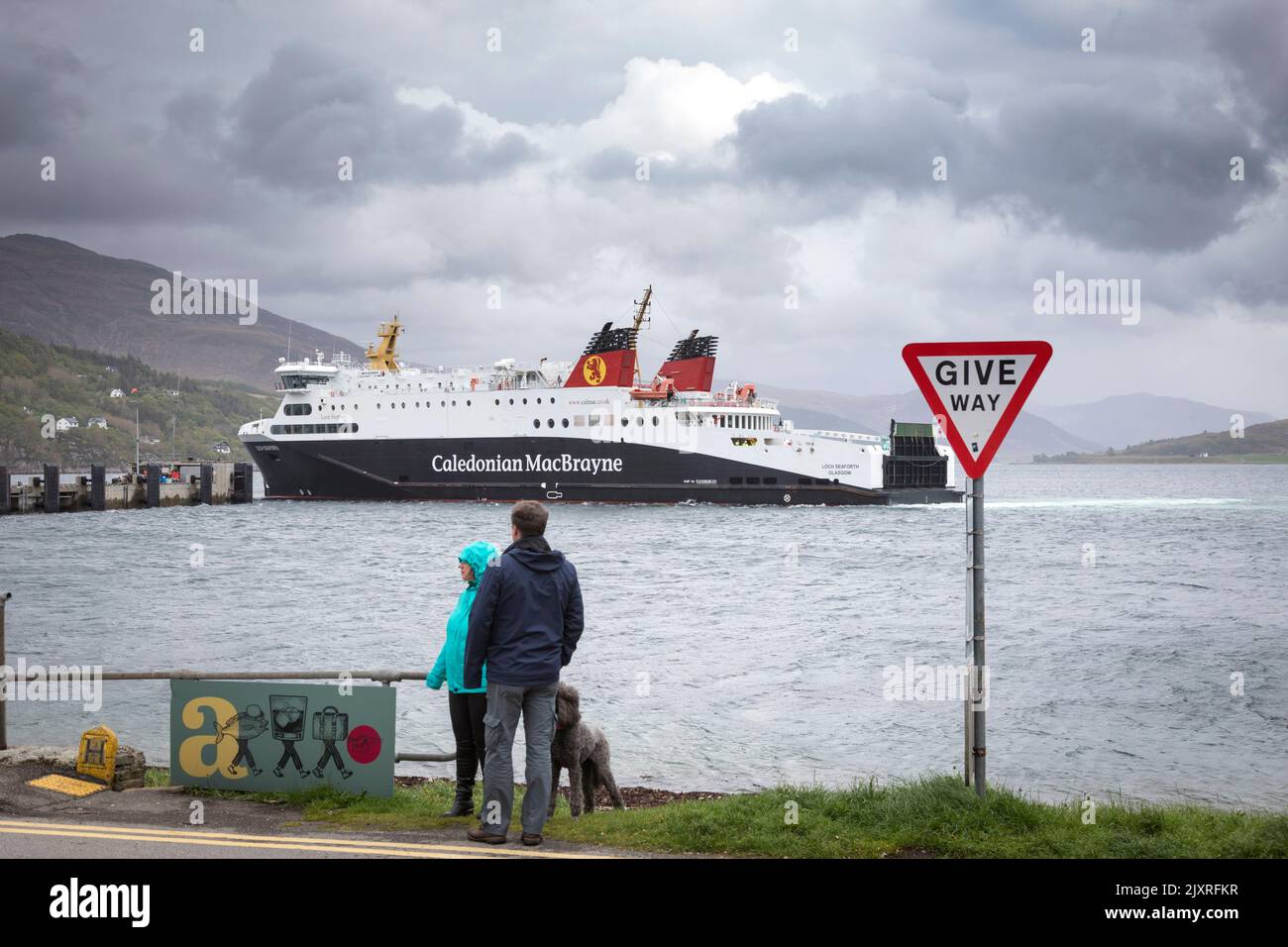 A couple walk their dog by the side of Loch Broom  as a CalMac ferry arrives at Ullapool harbour, Scotland. Stock Photo