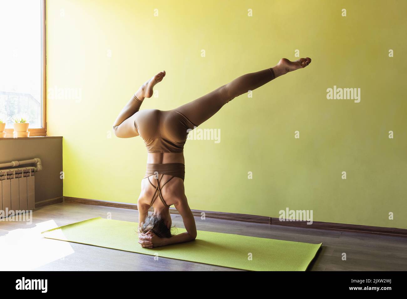 Young woman practicing yoga, performs a variation of the shirshasana exercise with a bent leg, inverted asana, stands on her forearms in the studio ne Stock Photo