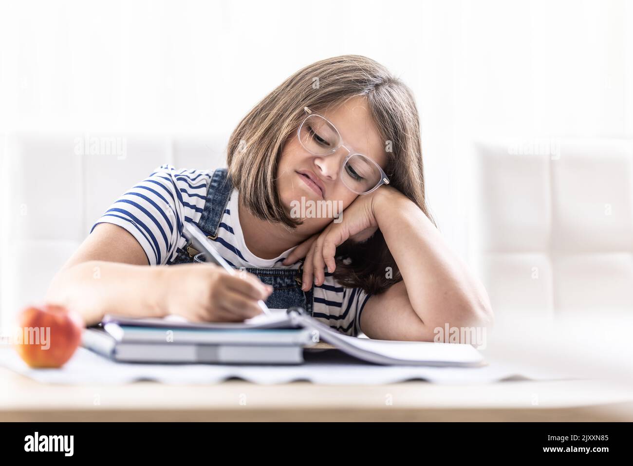 Annoyed schoolgirl with eyewear writes homework leaning against her hand. Stock Photo
