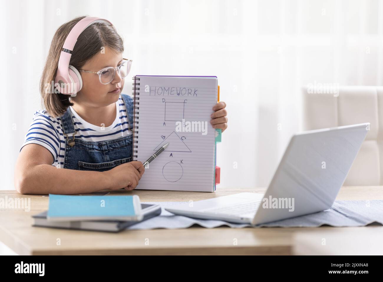 Schoolgirl in glasses and headphones shows her homework to the teacher during an online class via computer. Stock Photo
