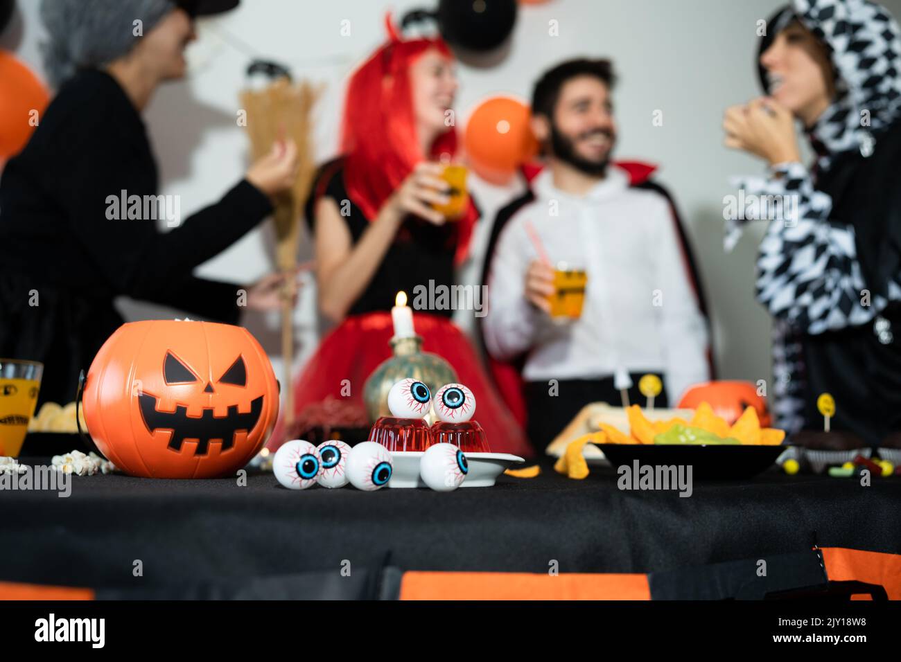 Group of friends in a halloween party. Devil, witch, dracula and harlequin eating, drinking and laughing Stock Photo