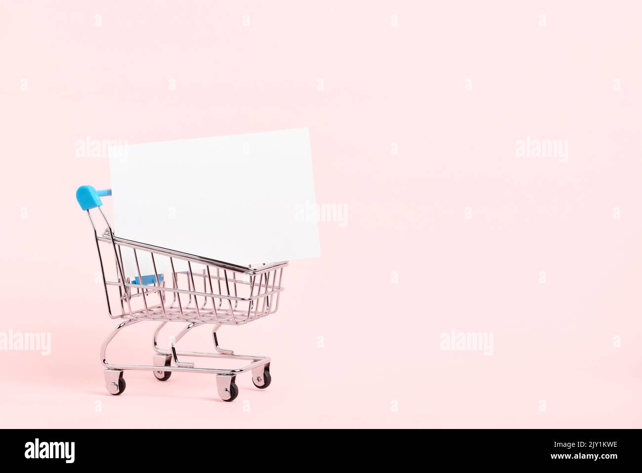 Shopping cart with a blank white paper card on a pastel pink background. Minimalist design with copy space. Concepts: market deals, seasonal sales and Stock Photo