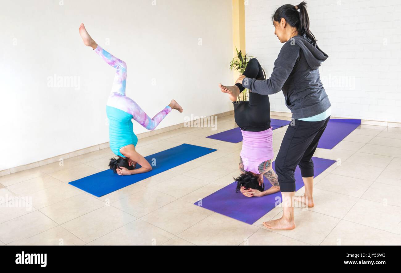 An instructor helps two women achieve headstand yoga posture with one leg twisted. Parivritta eka pada sirsasana Stock Photo