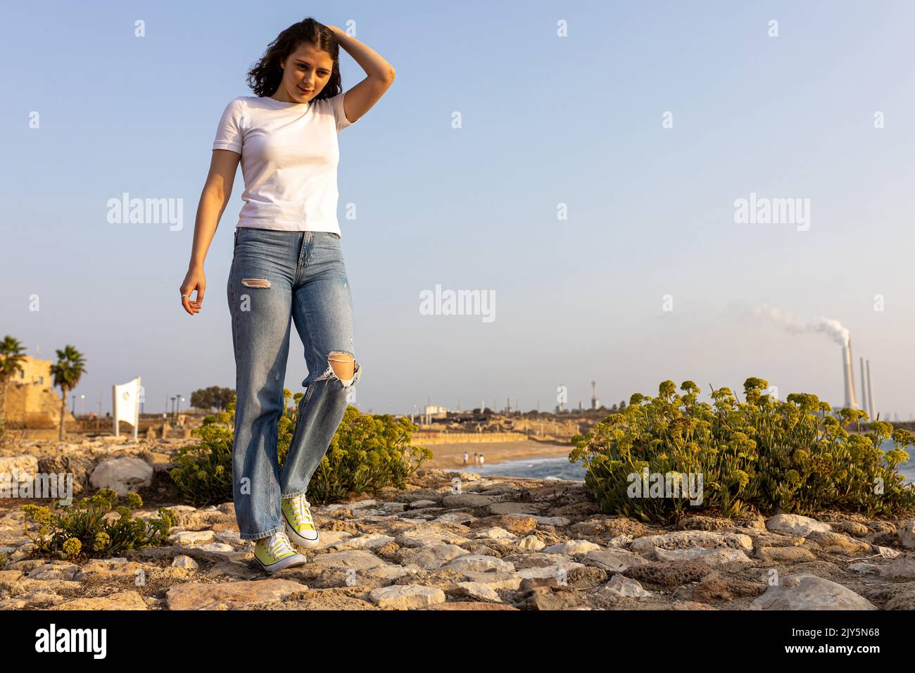 A smiling thoughtful girl in a white T-shirt, blue jeans and light green sneakers walks along the sea, balancing Stock Photo
