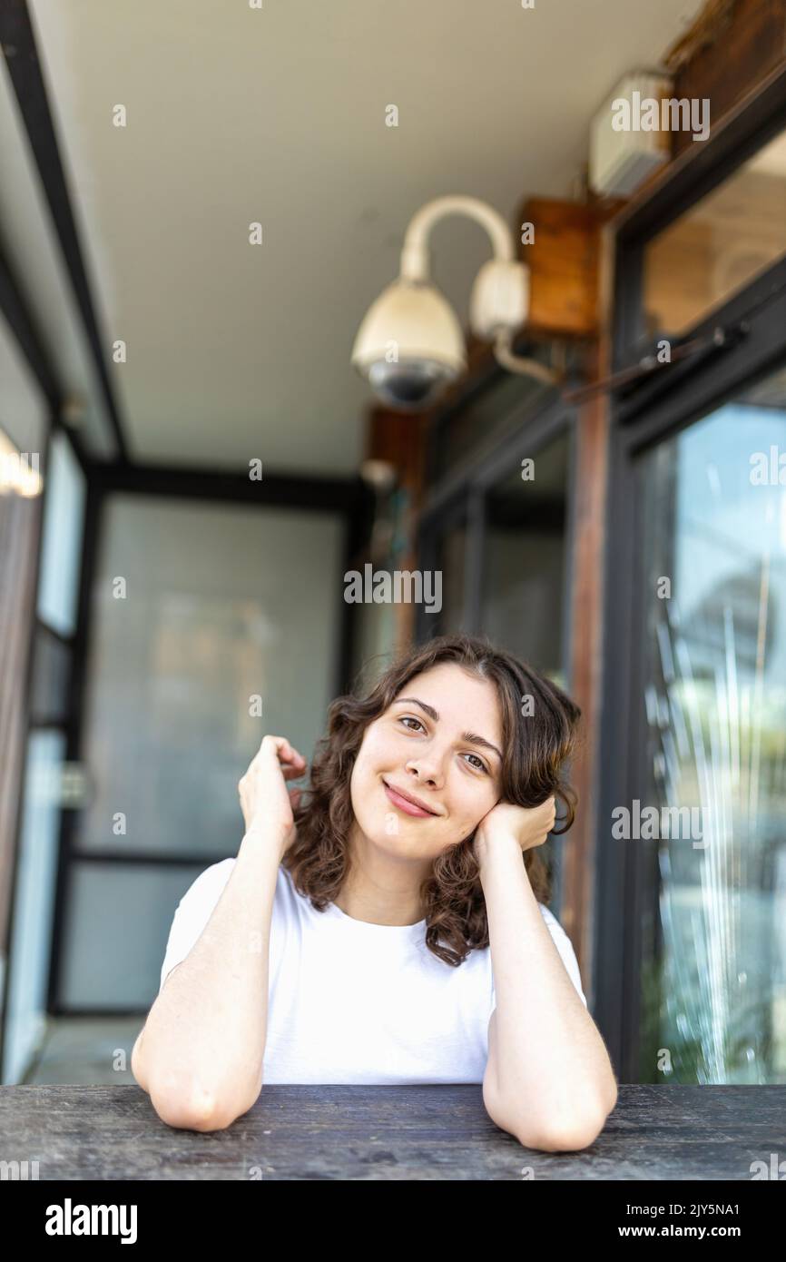 A smiling brunette girl in a white T-shirt, blue jeans sits at a wooden counter in a cafe, resting her chin in her hands. Vertical frame. Stock Photo